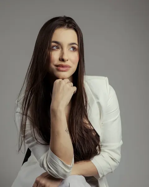 Tainá Cardoso sentada, de blazer branco, com a mão no queixo